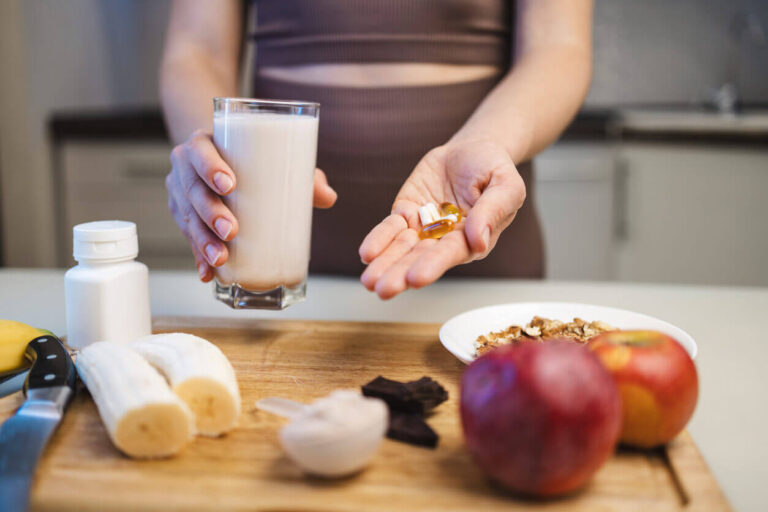 Pessoa segurando cápsulas de suplementos para colesterol ao lado de um copo de leite, frutas e cereais sobre a mesa, representando cuidados com a saúde