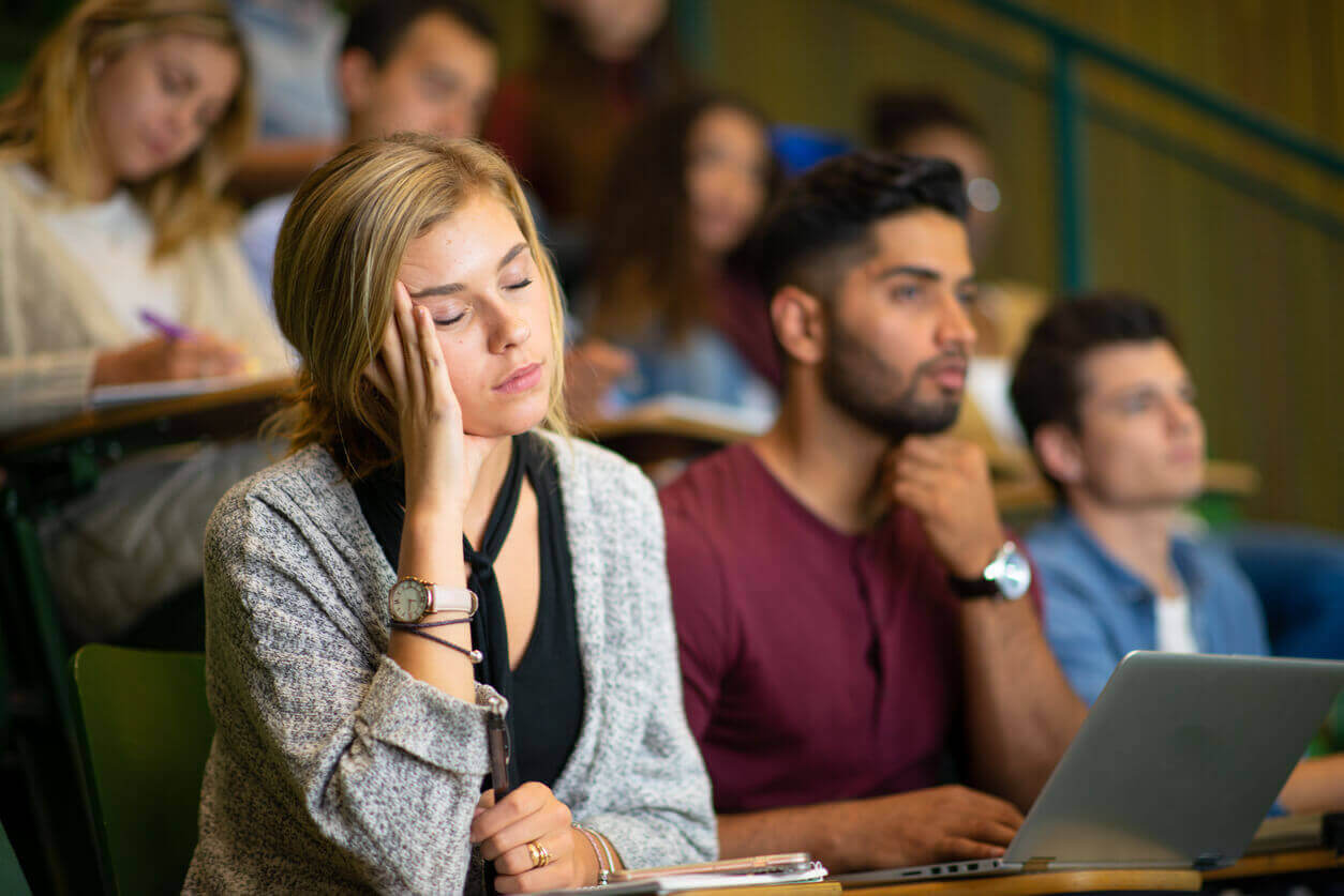 Mulher distraída em sala de aula enquanto colegas assistem à aula, cena que representa sinais de TDAH em mulheres no ambiente de estudo.
